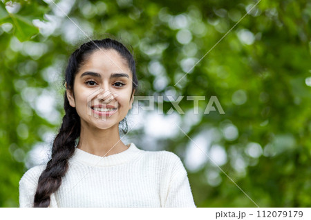 Close-up portrait of a young smiling Indian girl looking at the camera, standing in the park, outdoors. Close-up portrait of a young smiling Indian girl looking at the camera, standing in the park, outdoors. 112079179