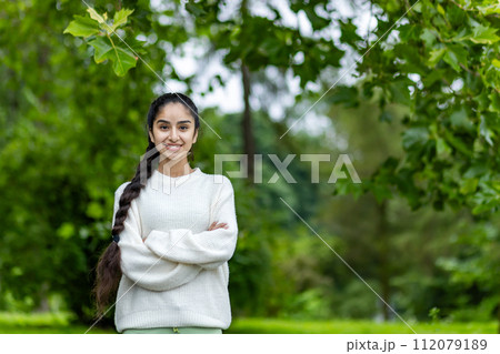 Portrait of a smiling Indian young girl in a white sweater doing sports and walking in the park. He stands with his hands crossed on his chest and looks confidently into the camera. 112079189