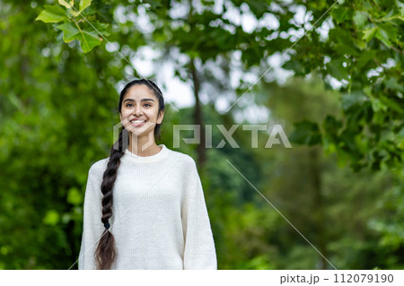 Photo of a beautiful young sporty Indian woman standing in a park in a white sweater and with a braid on her head, smiling and looking to the side. 112079190