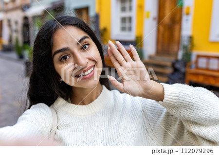 A beautiful young Indian woman in a white sweater is standing on a city street and talking on a video call on a telephone, waving and saying hello. A beautiful young Indian woman in a white sweater is standing on a city street and talking on a video call on a telephone, waving and saying hello. 112079296