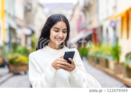 A young Indian girl walks on the city street and uses a mobile phone, calls, waits for an appointment, makes an online payment, rents in booking. Close-up photo. 112079304