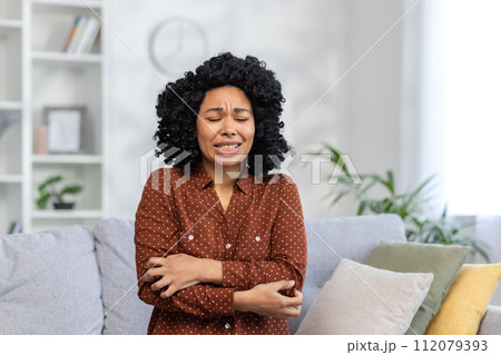 African American young woman sitting on the couch at home and shaking from the cold, having a panic attack and depression, wrapped her arms around herself and closed her eyes in fear. 112079393