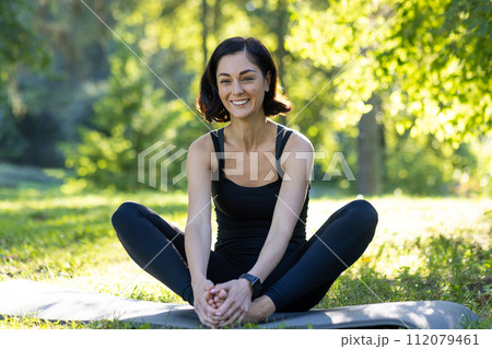 Content woman practicing mindfulness during outdoor yoga session in a tranquil park, surrounded by lush greenery. 112079461