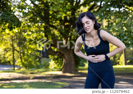 A young woman is standing in the park outdoors, bent over and holding her stomach, feeling severe pain after running and doing sports. 112079462