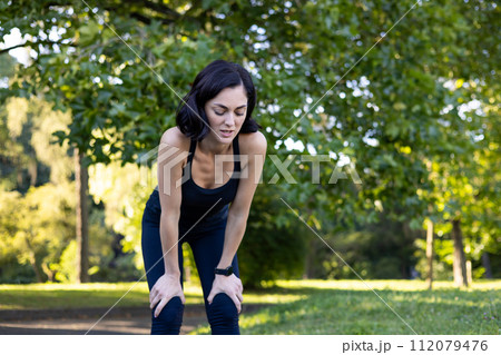 Tired young woman standing bent over in park, resting and shivering hard after jogging and training. 112079476