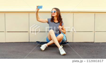 Modern happy smiling young woman taking selfie by smartphone sitting on skateboard on city street 112081817
