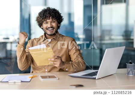 Rejoiced middle eastern male holding opened envelope and making winner gesture with hand in corporate building. Happy bearded businessman receiving mail letter with pleasant bank notification. Rejoiced middle eastern male holding opened envelope and making winner gesture with hand in corporate building. Happy bearded businessman receiving mail letter with pleasant bank notification. 112085216