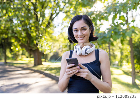 Close-up portrait of a smiling young woman doing sports in the park, going for a run, standing in headphones, holding a phone and looking at the camera. Close-up portrait of a smiling young woman doing sports in the park, going for a run, standing in headphones, holding a phone and looking at the camera. 112085235