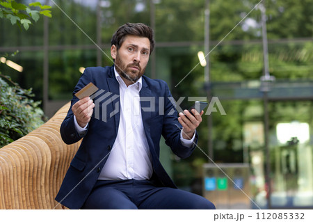 A businessman looks confused with a credit card and smartphone in hand, sitting outside an office, portraying finance issues. A businessman looks confused with a credit card and smartphone in hand, sitting outside an office, portraying finance issues. 112085332