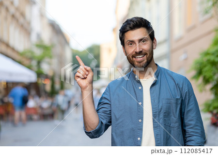 Portrait of a smiling young man standing on a city street in a blue shirt and pointing his hands to the side. Portrait of a smiling young man standing on a city street in a blue shirt and pointing his hands to the side. 112085417