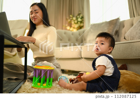 Cute little sitting on carpet near busy young beautiful mother working remotely on laptop Cute little sitting on carpet near busy young beautiful mother working remotely on laptop 112087125