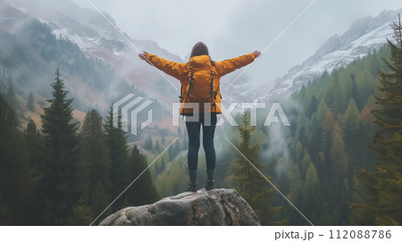 An adventurer in a bright yellow jacket stands with arms wide open, facing the misty mountainous landscape. An adventurer in a bright yellow jacket stands with arms wide open, facing the misty mountainous landscape. 112088786
