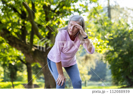 An active senior woman taking a break from running, appears tired, wiping sweat from her forehead in a peaceful park. 112088939