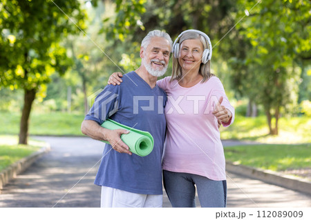 Cheerful senior man and woman in sportswear enjoying fitness lifestyle outdoors with yoga mat and headphones, showing thumbs up. Cheerful senior man and woman in sportswear enjoying fitness lifestyle outdoors with yoga mat and headphones, showing thumbs up. 112089009