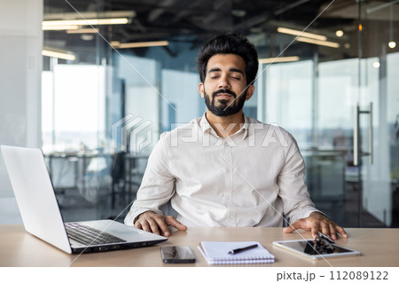 Relaxed young Indian man sitting in office at desk with laptop and documents and resting with closed eyes. Relaxed young Indian man sitting in office at desk with laptop and documents and resting with closed eyes. 112089122