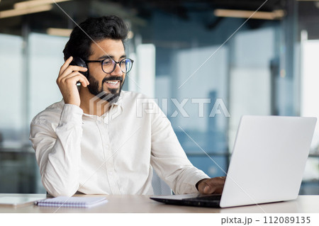 Indian young man businessman sitting in the office at the desk in front of the laptop and smilingly talking on the mobile phone. Indian young man businessman sitting in the office at the desk in front of the laptop and smilingly talking on the mobile phone. 112089135