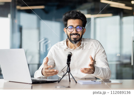 Smiling and successful young indian male businessman sitting in the office at the table in front of the microphone and having an online meeting, business training talking to the camera. Smiling and successful young indian male businessman sitting in the office at the table in front of the microphone and having an online meeting, business training talking to the camera. 112089136