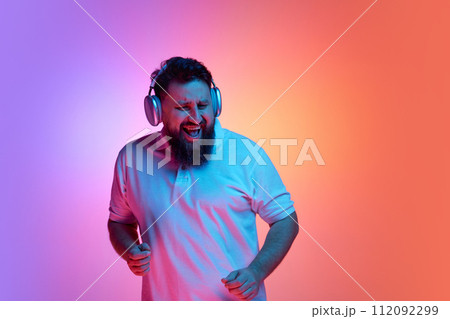 Overjoyed bearded man listening music in headphones and dancing in neon light against vibrant gradient studio background. Overjoyed bearded man listening music in headphones and dancing in neon light against vibrant gradient studio background. 112092299