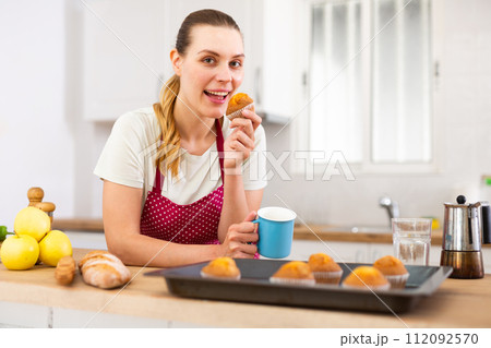 Smiling young woman in apron eating cupcakes from baking tray Smiling young woman in apron eating cupcakes from baking tray 112092570