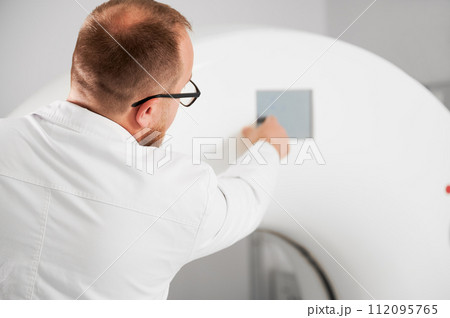 Medical computed tomography or MRI scanner. Back view of male radiologist presses MRI button to examine patient. Medical computed tomography or MRI scanner. Back view of male radiologist presses MRI button to examine patient. 112095765
