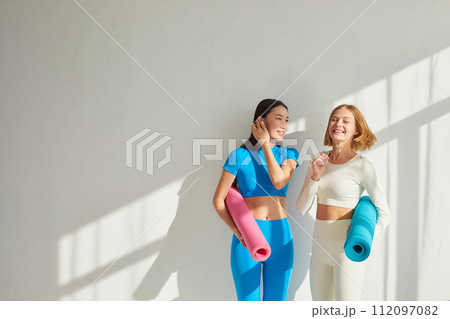 Two young women stand in yoga class, smiling and talking. Concept of physical and mental health care, asian and caucasian ethnicity 112097082