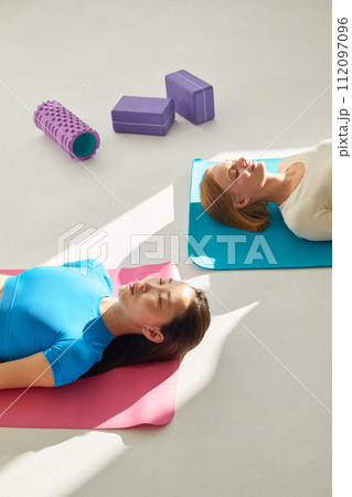 Two young women doing savasana pose after yoga class. Concept of physical and mental health care, deep meditation. Asian and caucasian ethnicity Two young women doing savasana pose after yoga class. Concept of physical and mental health care, deep meditation. Asian and caucasian ethnicity 112097096