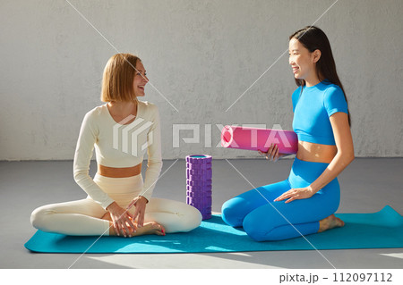 Two young women sitting in yoga class, smiling and talking. Concept of physical and mental health care, asian and caucasian ethnicity 112097112
