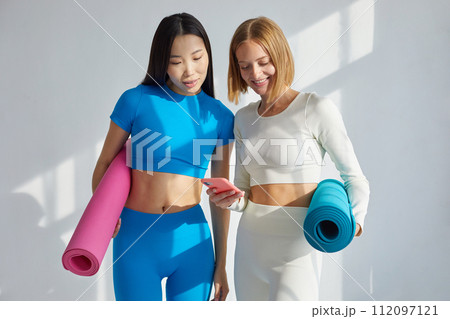 Two women watching yoga poses on smartphone while doing yoga. Concept of physical and mental health care, asian and caucasian ethnicity Two women watching yoga poses on smartphone while doing yoga. Concept of physical and mental health care, asian and caucasian ethnicity 112097121
