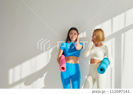 Two young women stand in yoga class, smiling and talking. Concept of physical and mental health care, asian and caucasian ethnicity 112097141