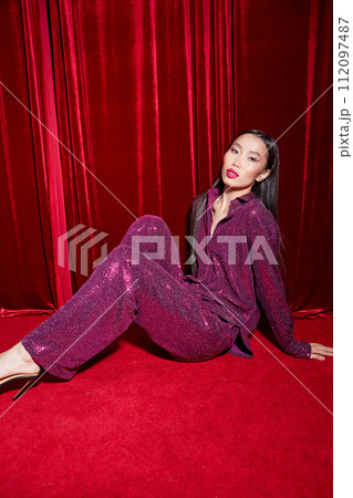 Fashion asian female model in burgundy glitter suit, on red fabric background. 112097487