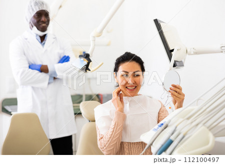 Satisfied asian woman looking in the mirror at the cured teeth at dentist Satisfied asian woman looking in the mirror at the cured teeth at dentist 112100497
