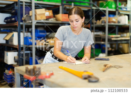 Young woman storehouse worker standing at workbench and making notes Young woman storehouse worker standing at workbench and making notes 112100533