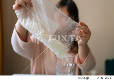Front view of unrecognizable female artisan pouring dry mixture or soy wax from transparent bag for creating candle building mixture. Process of making handmade natural scented candle at workshop. Front view of unrecognizable female artisan pouring dry mixture or soy wax from transparent bag for creating candle building mixture. Process of making handmade natural scented candle at workshop. 112101853