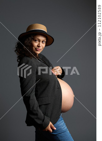 Attractive smiling happy pregnant woman holding her belly, dreamily looking aside a copy space on fashion gray backdrop 112102589