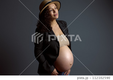 Attractive pensive young pregnant woman holding her belly, dreamily looking aside a copy space on fashion gray backdrop 112102590