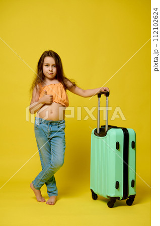 Little child traveler girl 6 years old in summer wear with suitcase, showing a thumb up over yellow studio background Little child traveler girl 6 years old in summer wear with suitcase, showing a thumb up over yellow studio background 112102624