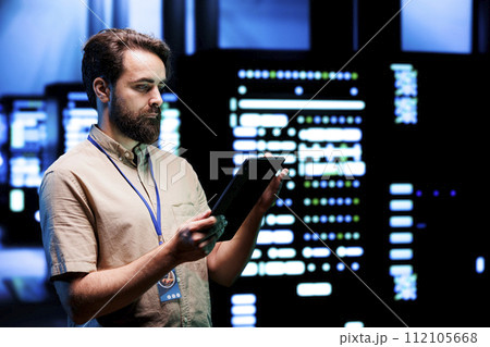 Worker doing system overhaul, imputing data on tablet of required patching in server cabinets. Facility housing advanced data storage equipment supporting critical IT workloads 112105668