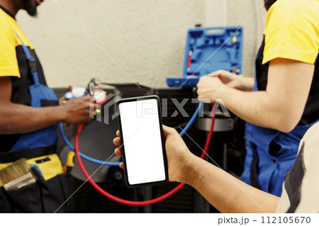 Capable specialist cleaning and lubricating external hvac system internal draining lines, checking freon level and calibrating thermostats while diverse team checks maintenance schedule on smartphone Capable specialist cleaning and lubricating external hvac system internal draining lines, checking freon level and calibrating thermostats while diverse team checks maintenance schedule on smartphone 112105670