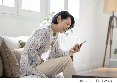下腹部をおさえながらスマートフォンを見るアラフィフの女性　腹痛　イメージ 112105691