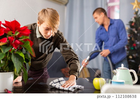 Son cleans the table with a rag while father vacuums room before christmas Son cleans the table with a rag while father vacuums room before christmas 112108638