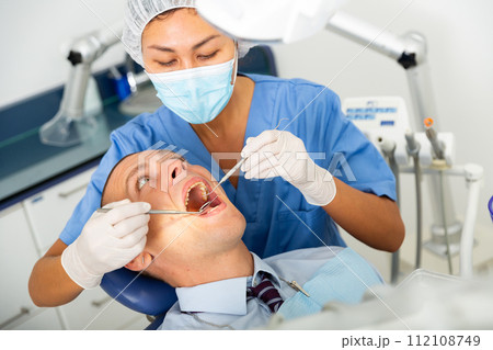 Qualified woman dentist in a protective mask treats a man patient Qualified woman dentist in a protective mask treats a man patient 112108749