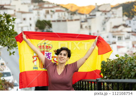 Cheerful young brunette standing outdoors with national flag of Spain 112108754