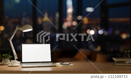 Office at night, a laptop computer mockup, a table lamp, and accessories on a wooden desk. 112109715