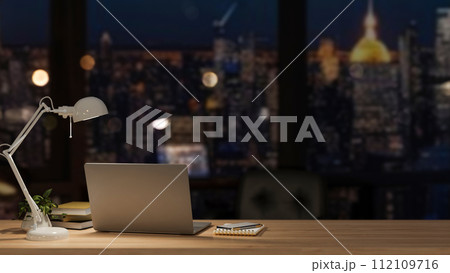 A modern office at night with a city view, featuring a laptop and accessories on a wooden desk. 112109716