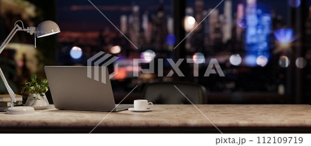 A modern office at night with a city view, featuring a laptop and accessories on a desk. 112109719