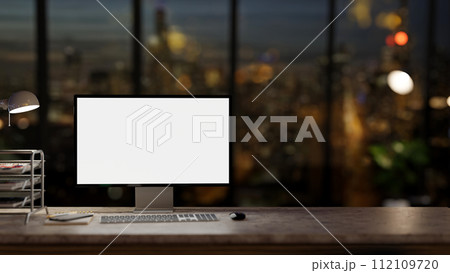 A computer desk in a modern dark office at night with a city view through the large glass window. 112109720