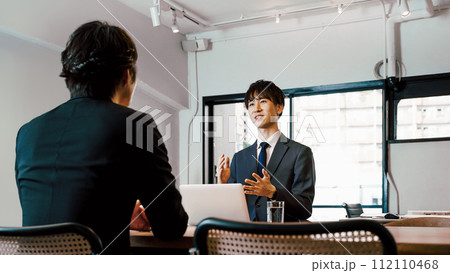 会議室のあるオフィスで対面で商談の打ち合わせをするスーツのビジネスマンの2人の男性 112110468