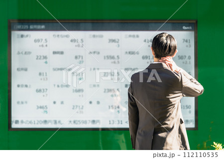 電光掲示板の株価ボードを見てその不景気に首に手を当てて落胆する30代のスーツ姿のビジネスマンの後ろ姿 112110535