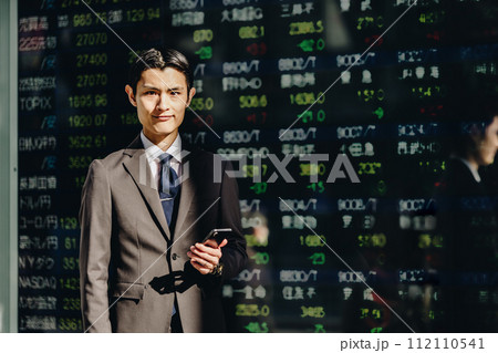 電光掲示板にうつる株価ボードの前でスマートフォンを持って立つカメラ目線の30代の男性 112110541