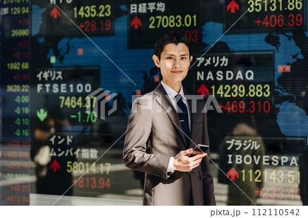 世界中の株価が映る電光掲示板の前で笑顔で立つカメラ目線の30代の男性（戦略のイメージ） 112110542